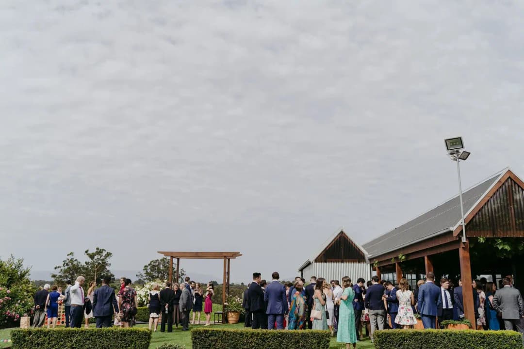 Guests gathered beside an outdoor ceremony arch near a timber reception building.
