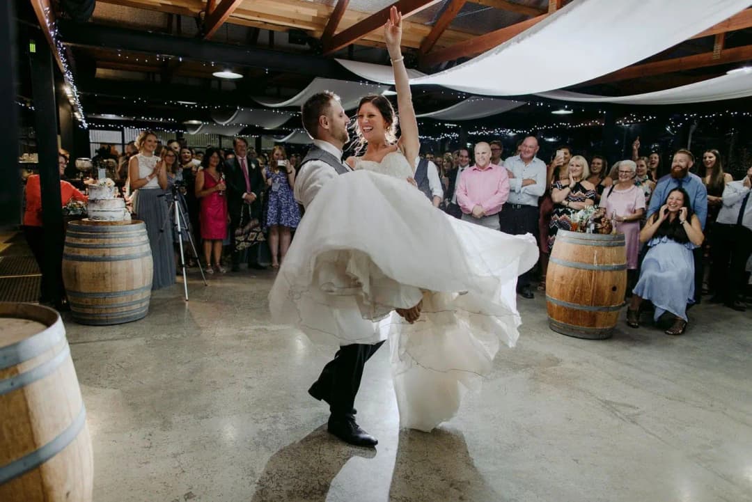 Groom lifting the bride across his arms while guests applaud on the dance floor.