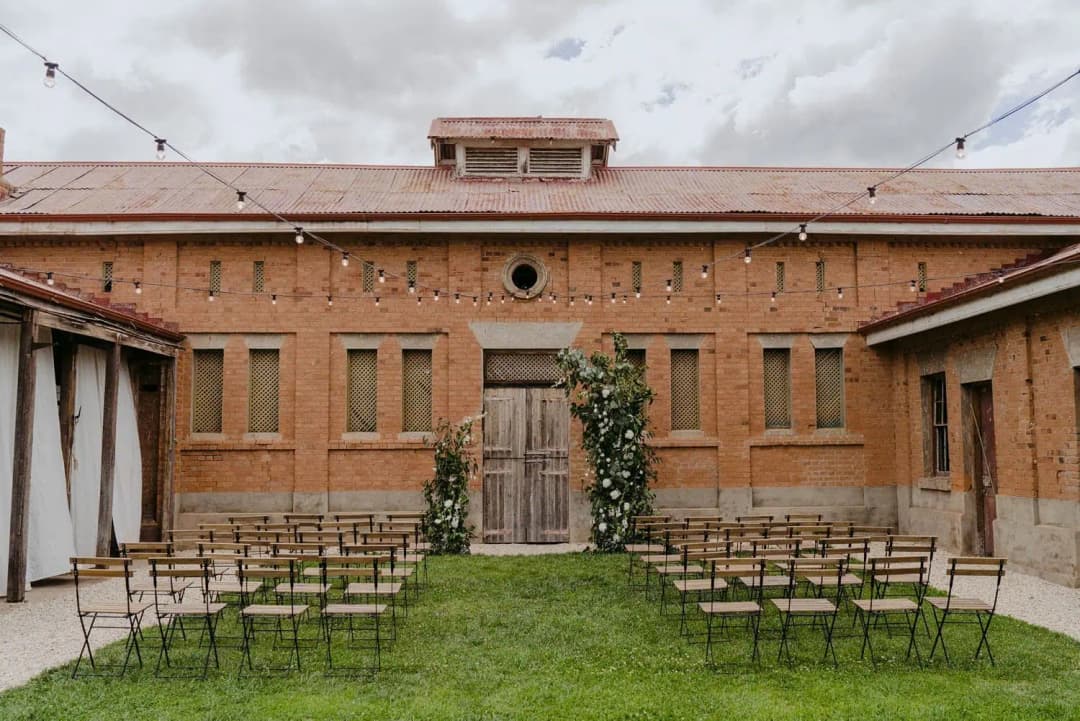 Rows of wooden ceremony chairs facing a floral arch outside a brick building.