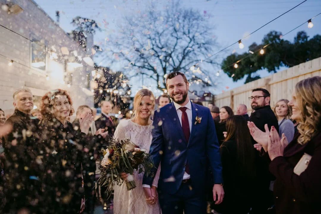 Bride and groom smiling through confetti thrown by guests in a courtyard.