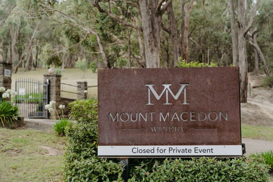 Rust-coloured winery sign beside garden plants and tall trees.