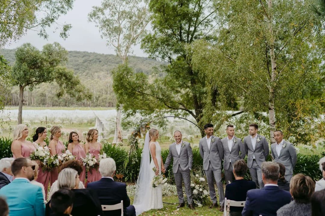 Wedding party facing guests at a garden ceremony beneath leafy trees.