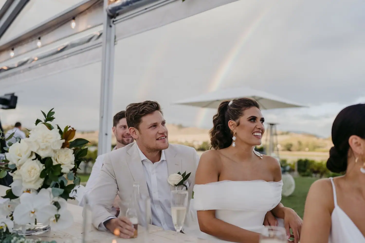 Bride and groom smiling at their reception table beneath a rainbow.