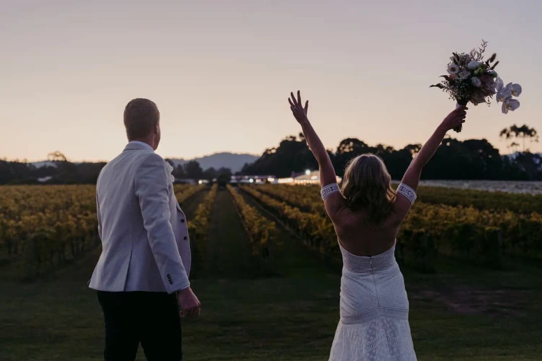 Bride raising her bouquet and both arms beside the groom at sunset.
