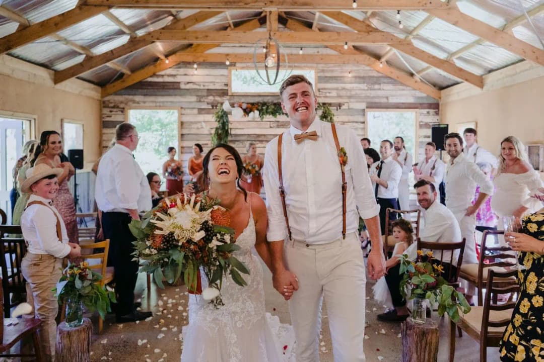 Bride and groom laughing as they walk between guests inside a timber-roof ceremony room.