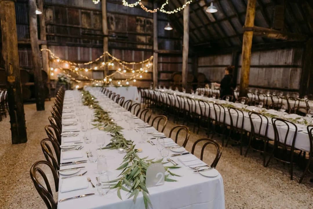 Greenery arranged along a long white table beneath warm lights in a rustic shed.