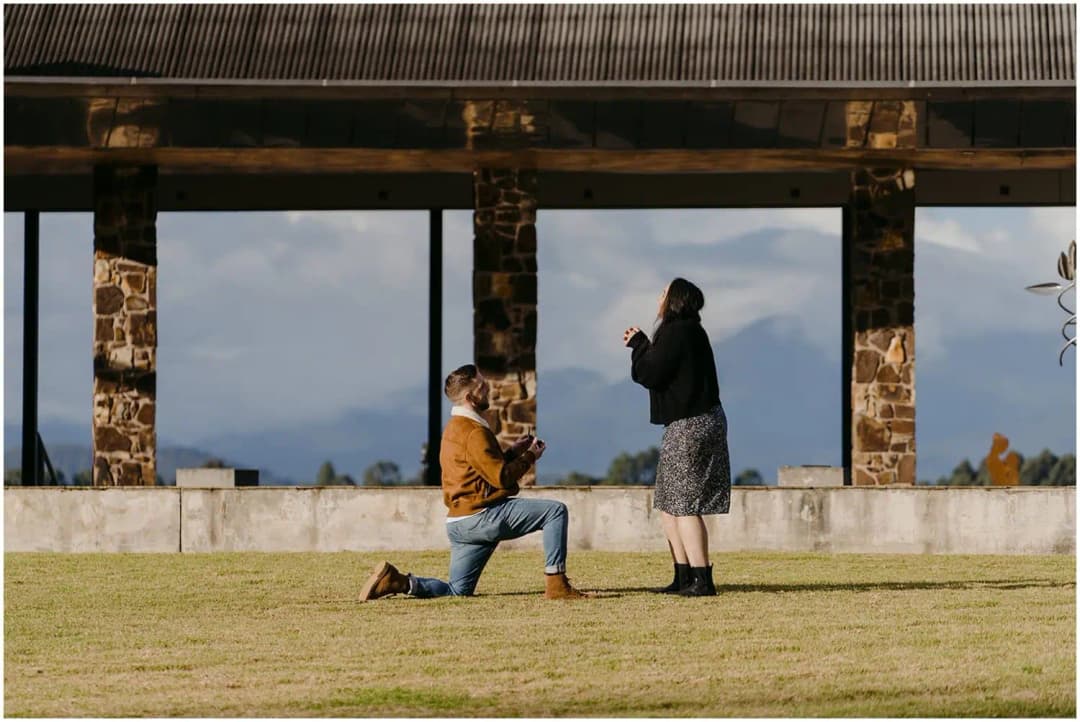 Man kneeling before a woman beside a roofed stone structure.