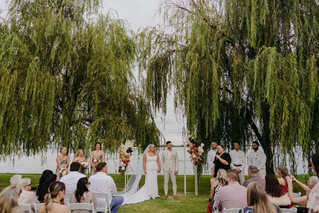 Wedding party and guests gathered beneath willow trees for a lakeside ceremony