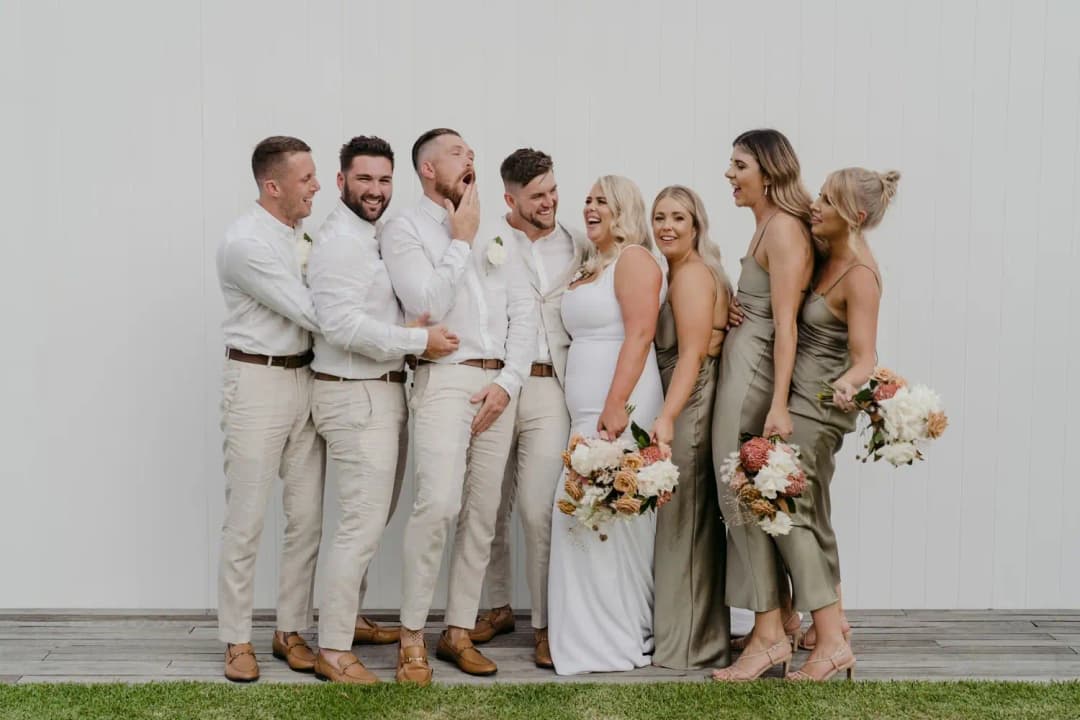 Bride and groom smiling with their wedding party beside a white wall