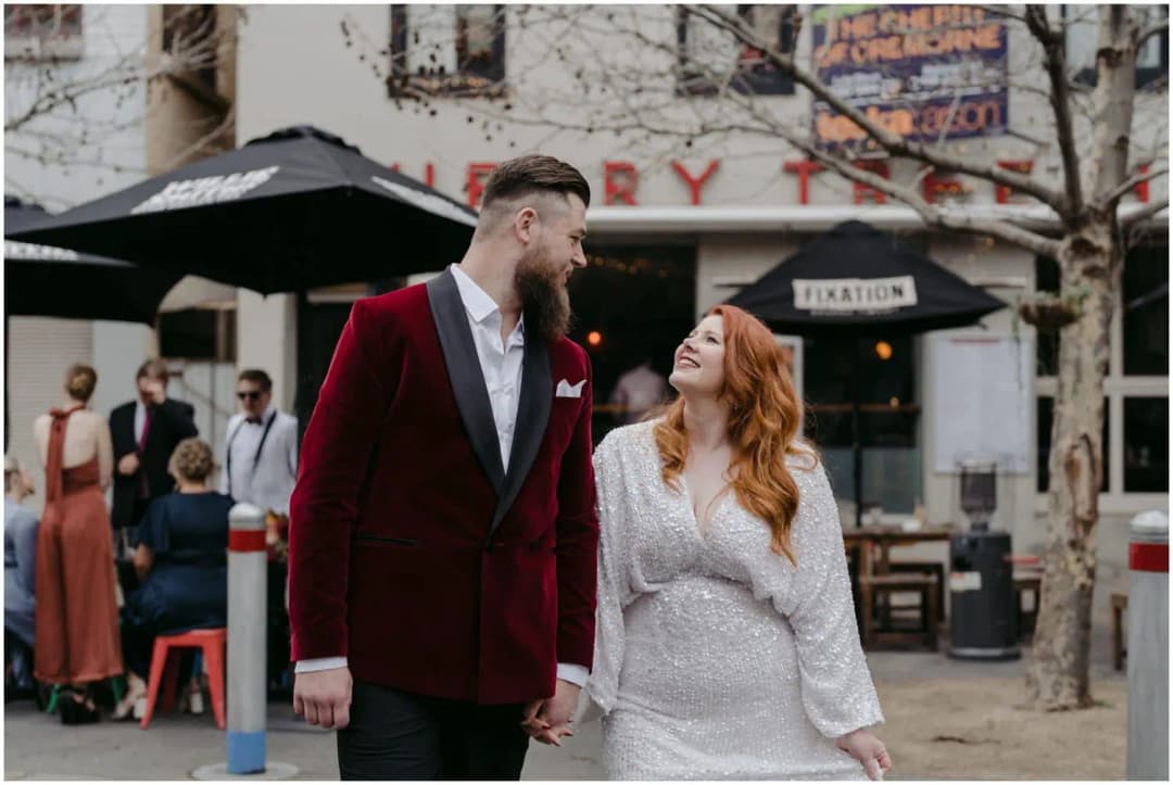 Bride and groom holding hands and smiling while walking past a pub