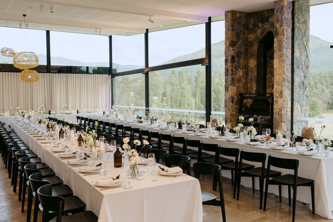 Long white reception tables beside a tall stone fireplace and wide windows.