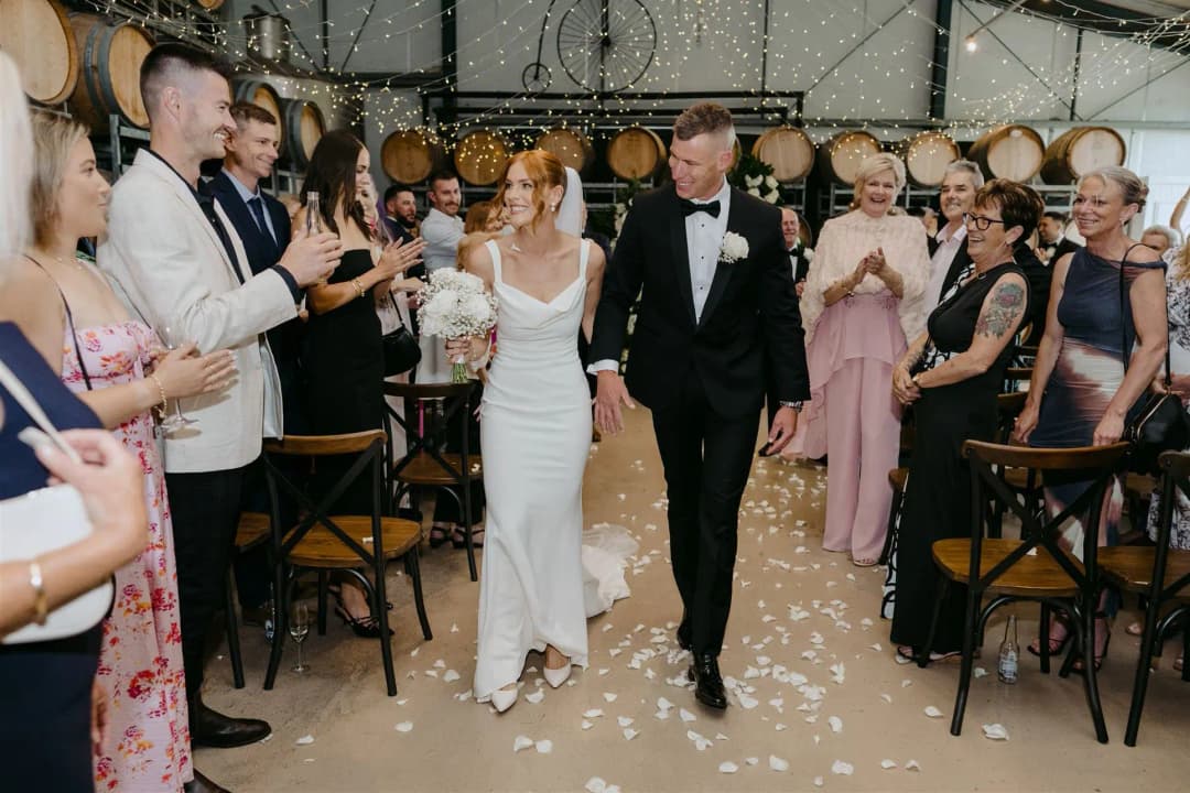 Couple walking down an indoor aisle through white petals and applauding guests.