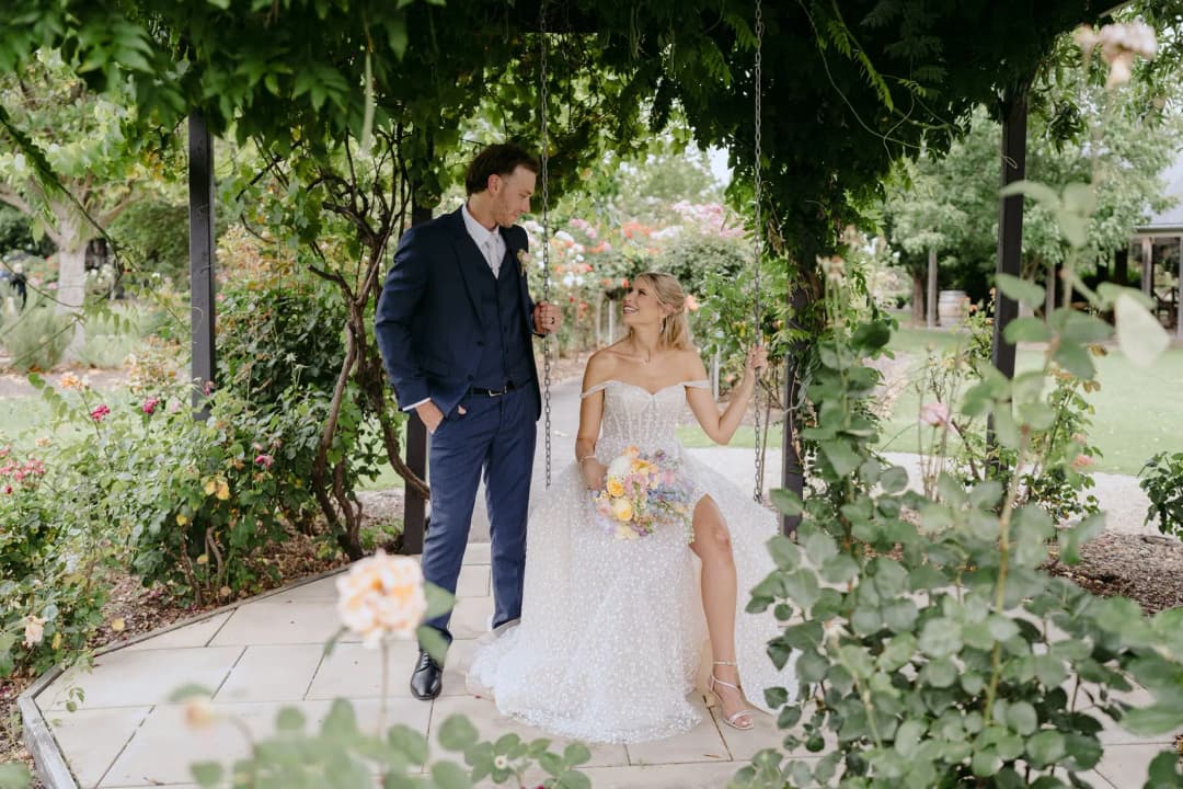 Bride seated beside the groom beneath a leafy garden pergola.