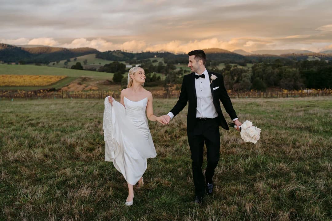 Couple walking hand in hand across a field beneath sunset-lit mountains.