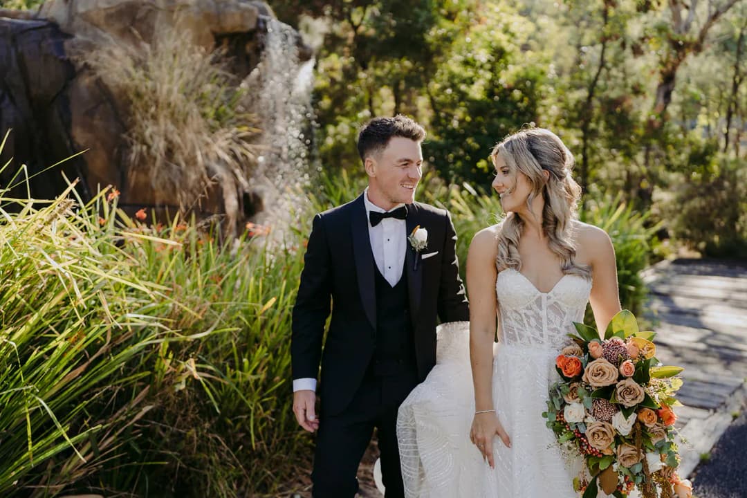 Bride and groom smiling at each other on a leafy garden path.