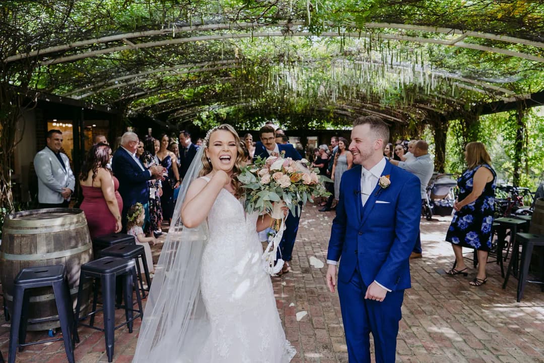 Couple laughing and walking through guests beneath a vine-covered pergola.