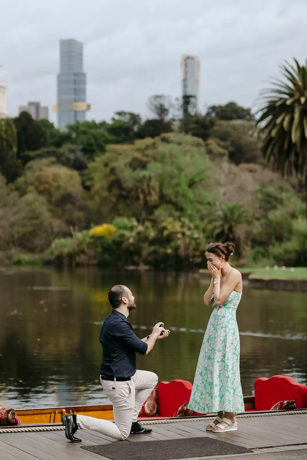 Man kneeling before a surprised woman beside a lake and small boat.