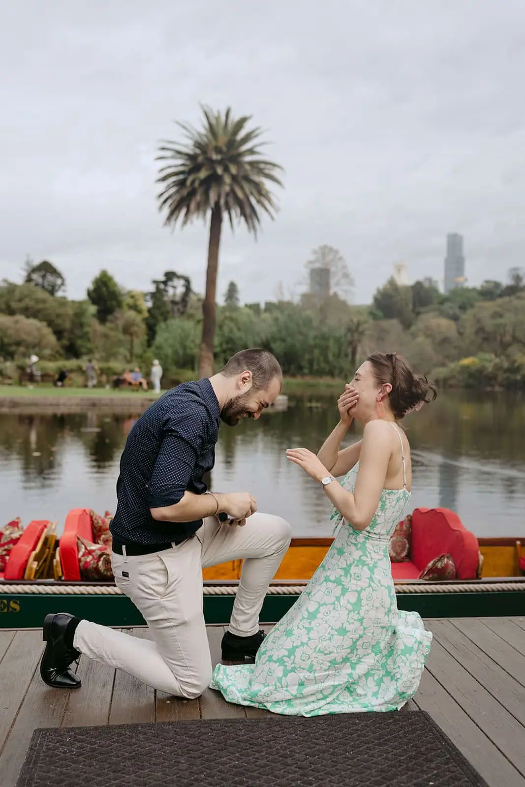 Woman covering her mouth as a man kneels beside her on a lakeside dock.