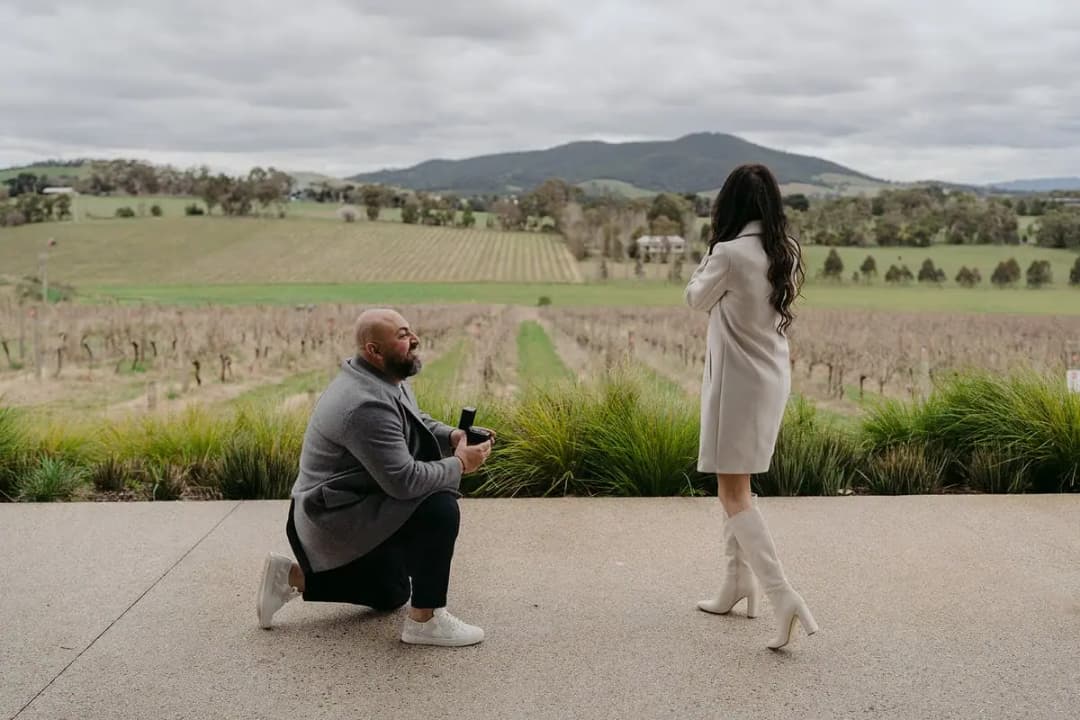 Woman covering her mouth as a man kneels before her on a terrace.