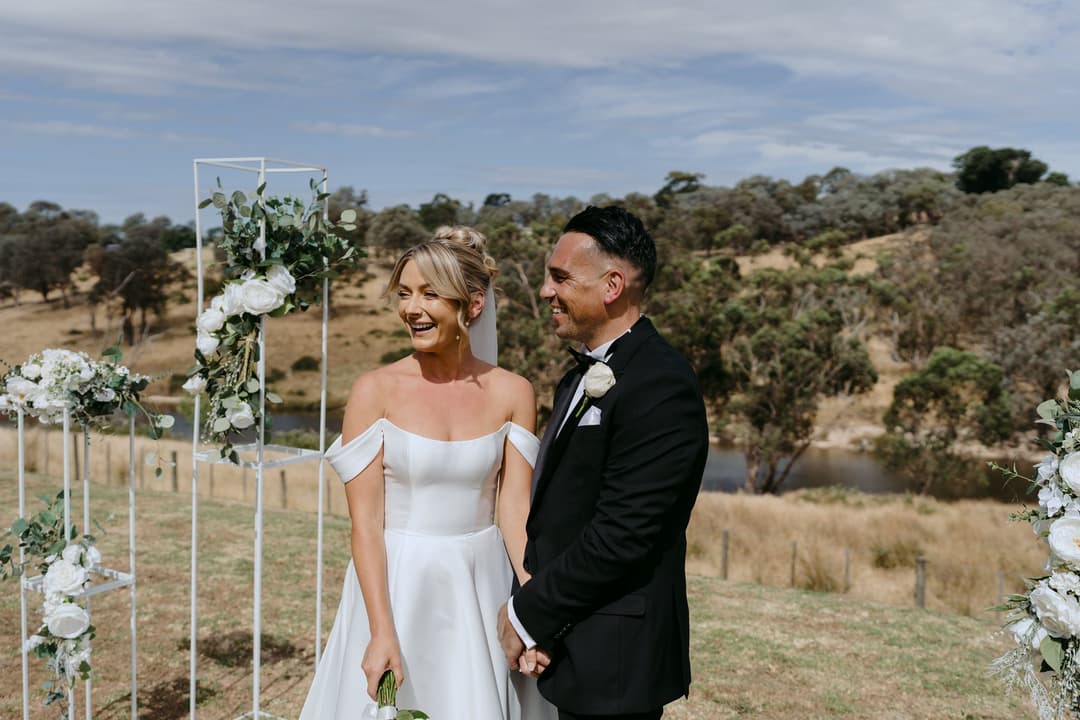Couple smiling beside a white floral ceremony arch overlooking hills.