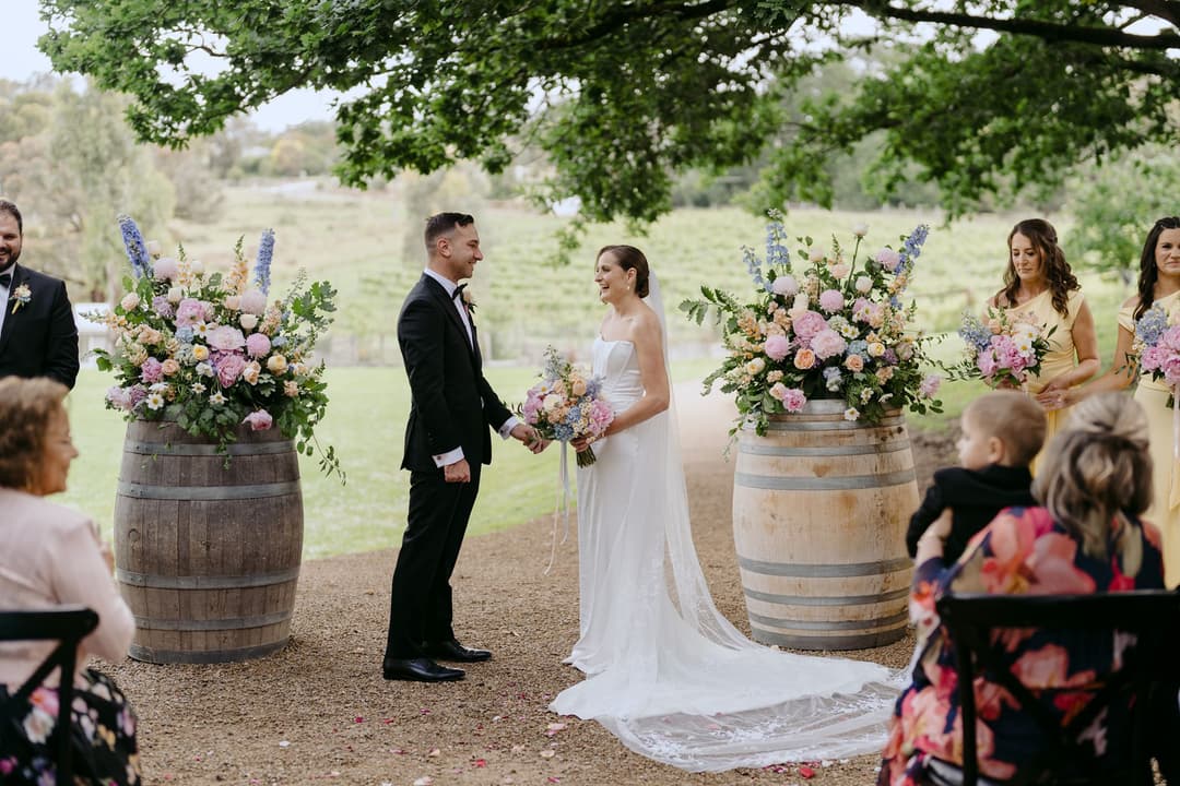 Couple facing each other between flower-topped barrels at a garden ceremony.