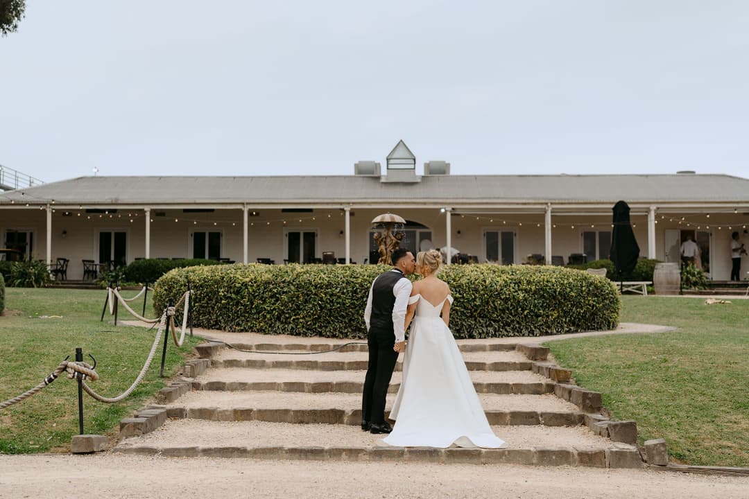 Couple embracing on stone steps outside a long building with a veranda.